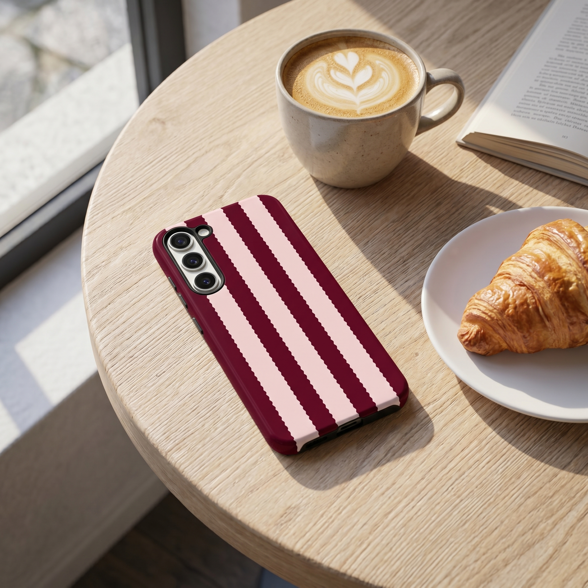 Phone case with striped burgundy and pink design on a table with coffee and croissant.