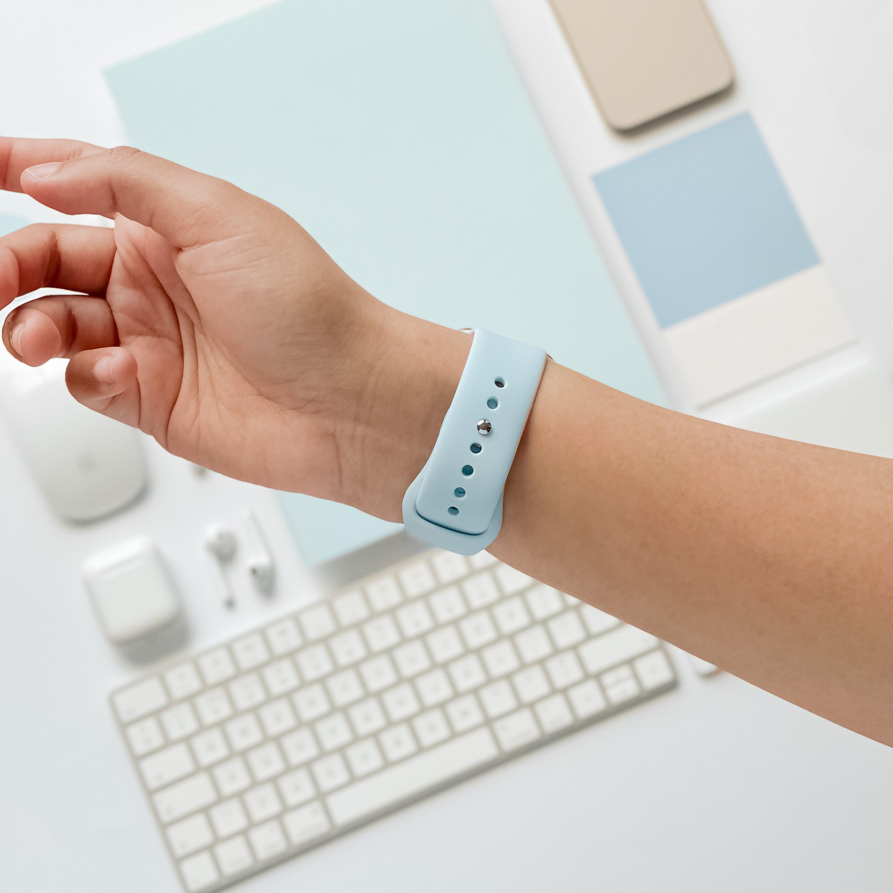 Close-up of a person’s left wrist wearing a Coconut Lane NAKD Apple Watch Strap in Sky Blue. In the background, there’s a wireless mouse, keyboard, earbuds, phone, and other spring-inspired pastel tech accessories arranged neatly on a white surface.