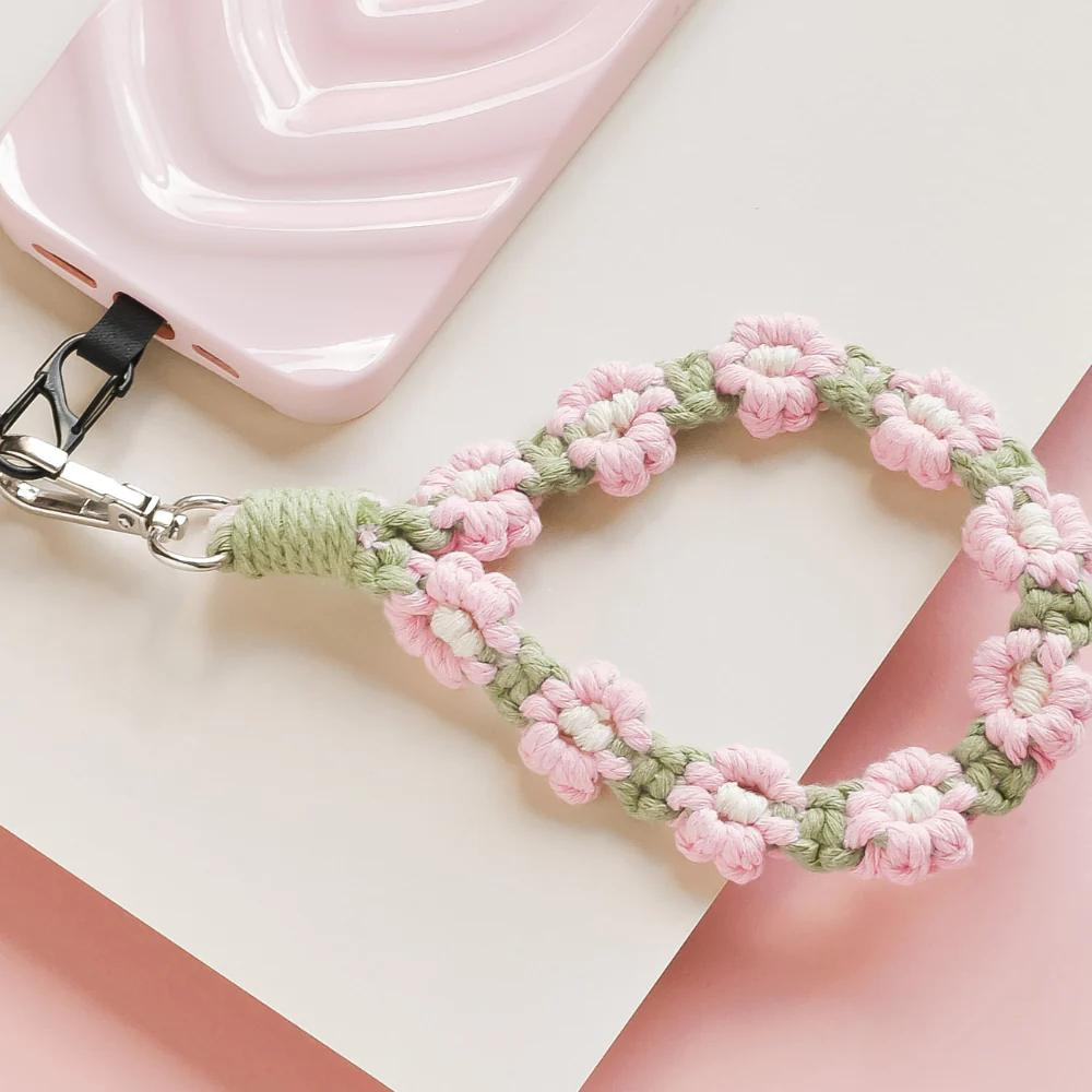 A pink and green handmade crochet phone strap featuring flower designs is attached to a pink phone case. The phone and strap are placed on a light pink and white background.