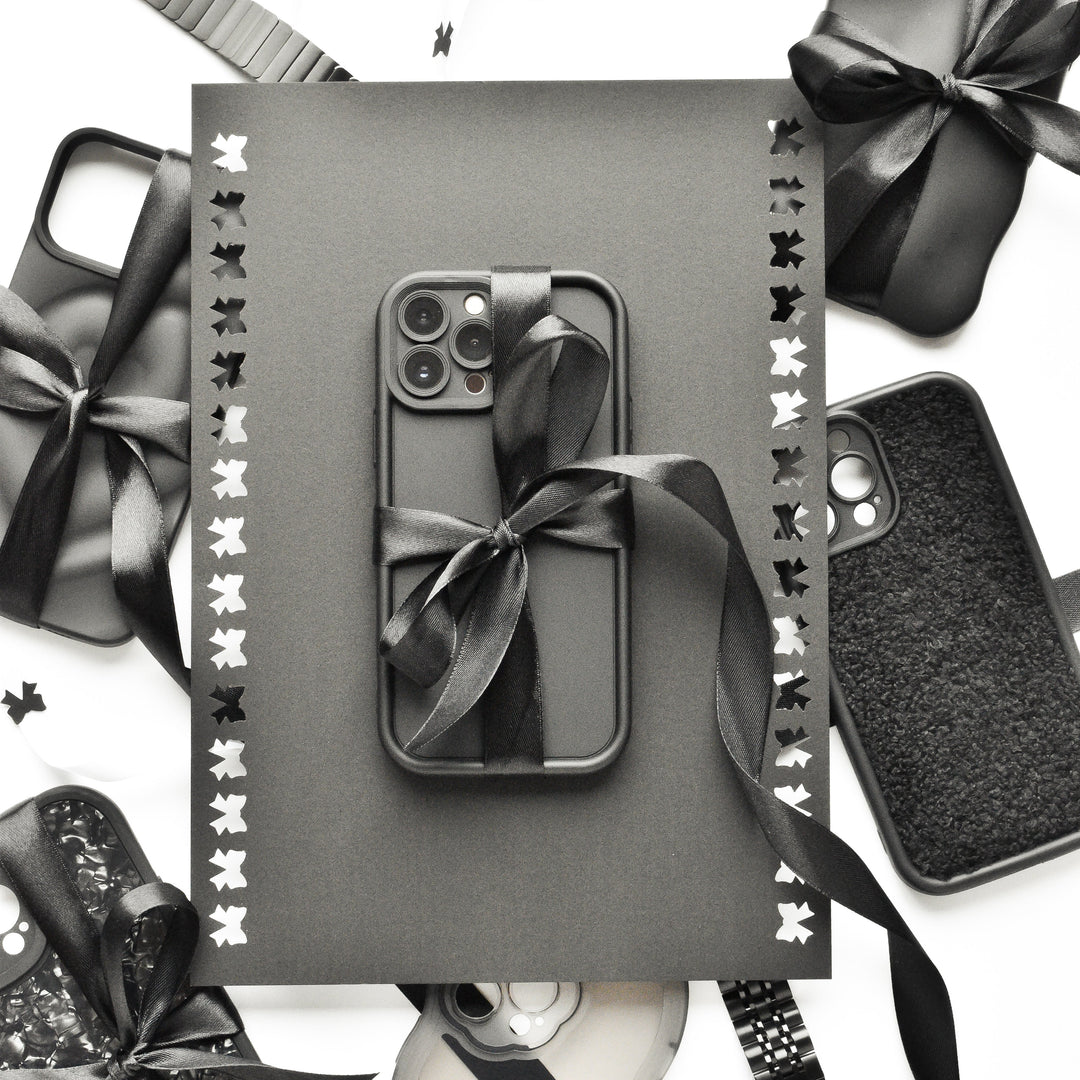 A black smartphone case with a ribbon lies on a black card with cut-out butterflies, surrounded by various black phone cases and accessories on a white background.