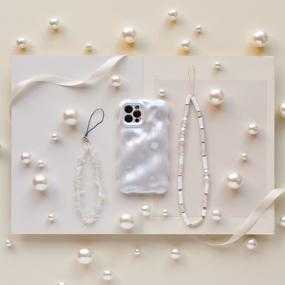 A pearl-themed flat lay featuring a textured phone case, two beaded phone charms on white open pages, and scattered pearls with a ribbon across a light beige background.