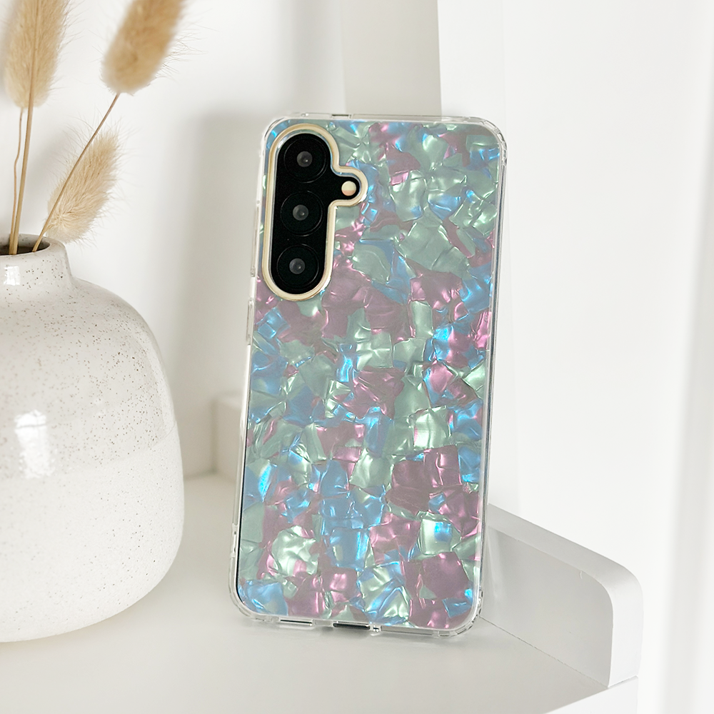 A smartphone in a colorful, iridescent case with a pattern resembling pieces of colorful foil sits upright on a white surface. A white vase with beige ornamental grass is in the background on the left.