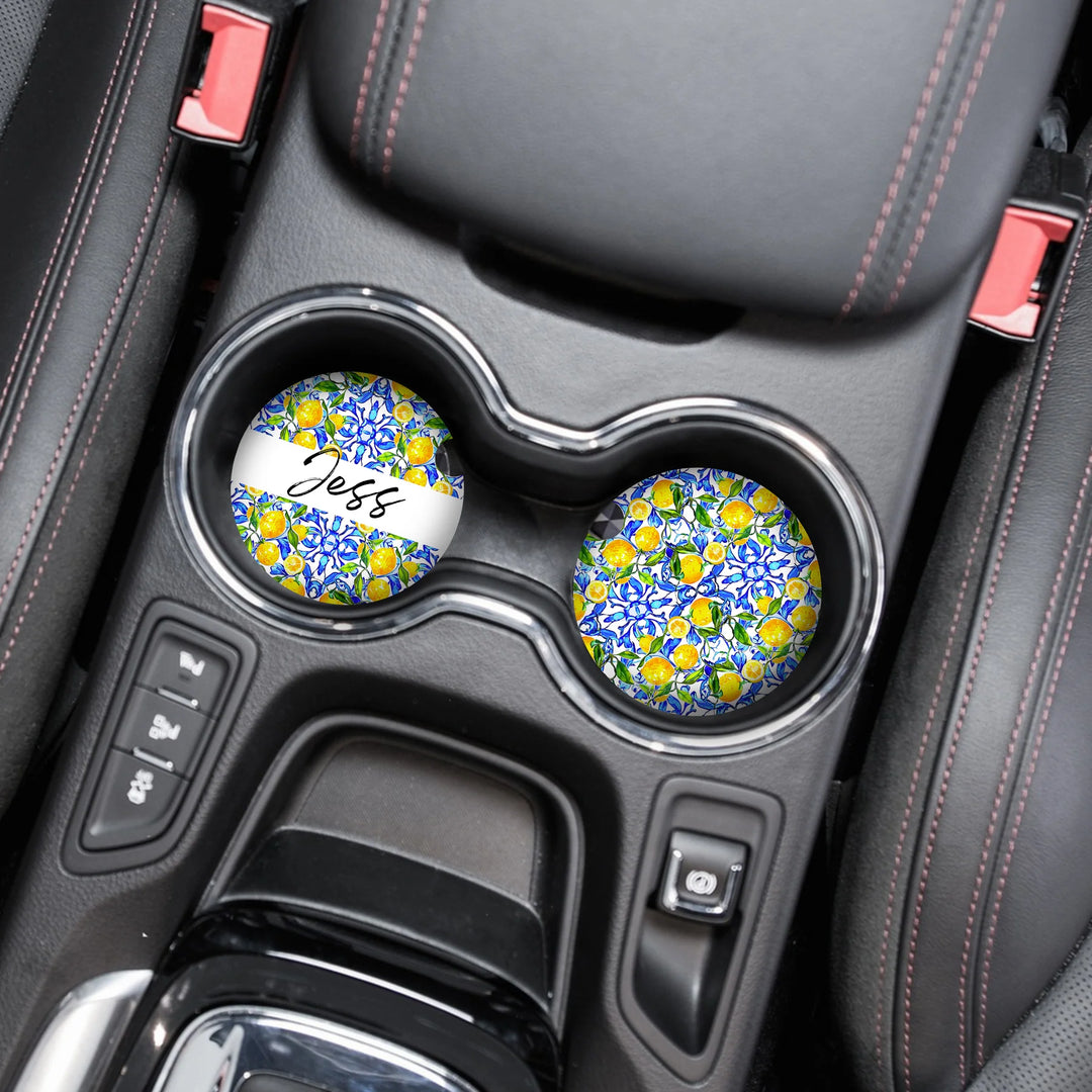 Automobile cup holders with decorative coasters featuring a vibrant blue, yellow, and green floral pattern. One coaster has the name "Jess" written on it. The car interior has black leather seats and red stitching.