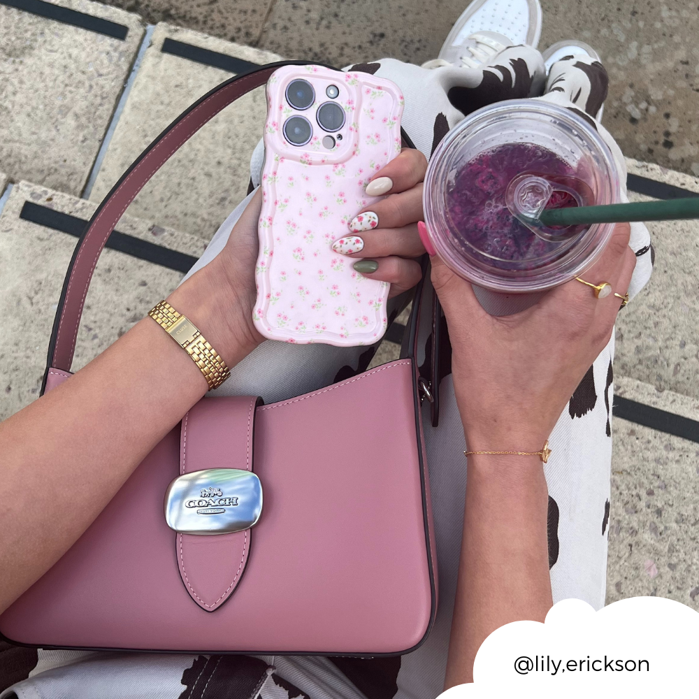 Person holding a Coconut Lane Wavy Phone Case - Ditsy Floral Pink and a purple drink in a clear cup. They have manicured nails and wear a gold watch, bracelet, and rings. A pink Coach handbag rests on their lap. The background is blurred, focusing on accessories. Instagram handle @lily.erickson.