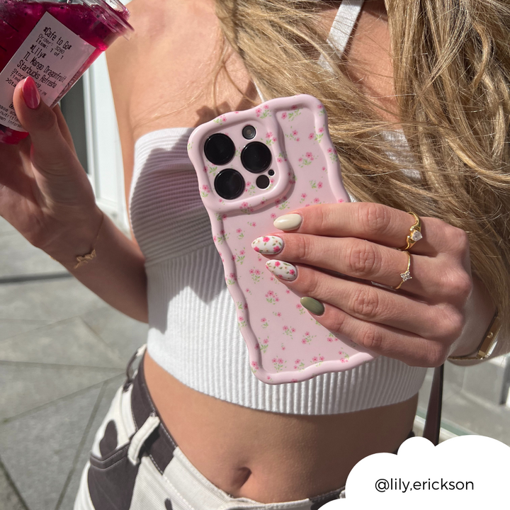 A person with long blonde hair is holding a phone adorned with Coconut Lane's Wavy Phone Case - Ditsy Floral Pink in one hand and a jar of a pink beverage in the other. They are wearing a white crop top and have pastel, floral-patterned nails. Their gold jewelry includes a ring and bracelet. The username "@lily.erickson" is displayed in the bottom-right corner.