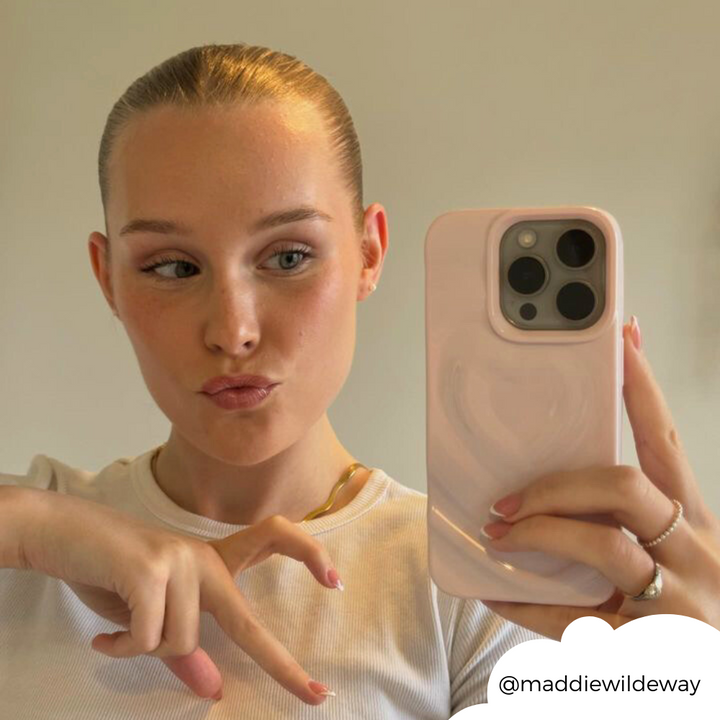 A person with slicked-back hair is taking a mirror selfie with a Melting Heart Phone Case in Peony Pink from Coconut Lane. They are wearing a white top and making a playful pouting expression. The Instagram handle @maddiewildeway is displayed at the bottom right with a cloud-like border.