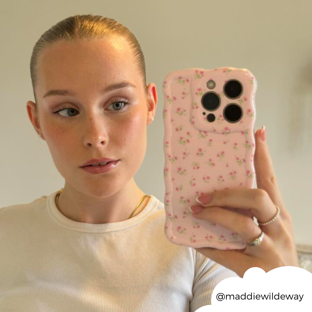 A person with their blonde hair tied back takes a mirror selfie. They are wearing a white top and holding a phone with the Wavy Phone Case - Ditsy Floral Pink by Coconut Lane. The Instagram handle @maddiewildeway is visible in the bottom right corner.