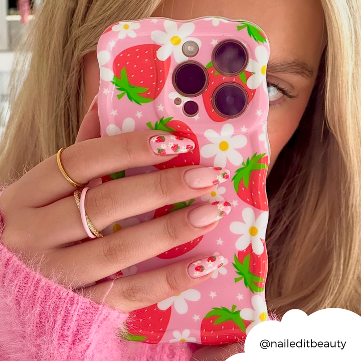 Close-up of a person holding a phone with Coconut Lane's Wavy Phone Case - Berry Cute Strawberry, featuring an adorable strawberry and daisy-themed design. The phone partially covers their face, perfectly matching their strawberry and floral nail art. They are wearing a pink sweater and gold rings. "@naileditbeauty" is displayed in a cloud shape.