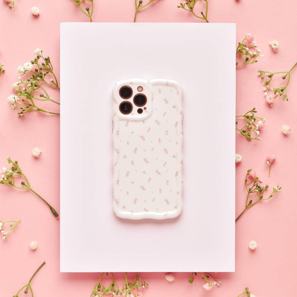 A smartphone encased in Coconut Lane's Wavy Phone Case - Ditsy Floral White lies on a white rectangular sheet, surrounded by delicate sprigs of small white flowers on a pink background. The overall aesthetic is soft and feminine.