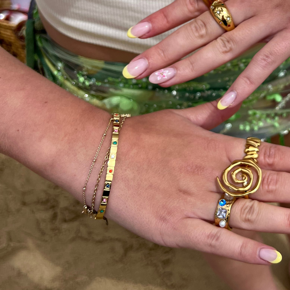 Hand with pastel yellow French-tip nails and gold rings, including a Sunset Spiral Ring, on green sequins.