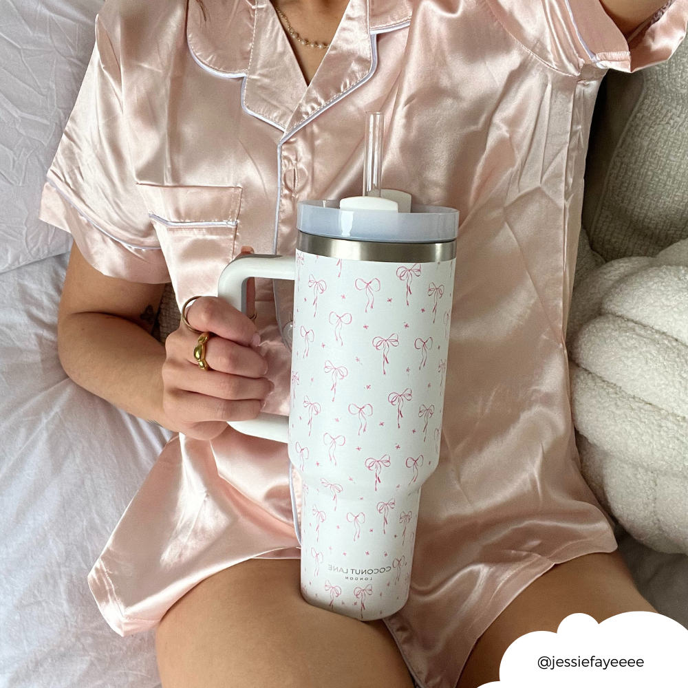 Person in pink satin pajamas holding a Coconut Lane stainless steel tumbler with dainty bows and reusable straw.