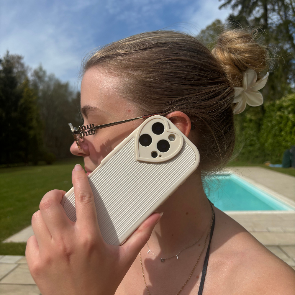 Woman by a pool holding a cream ribbed heart-shaped phone case with lens protection.