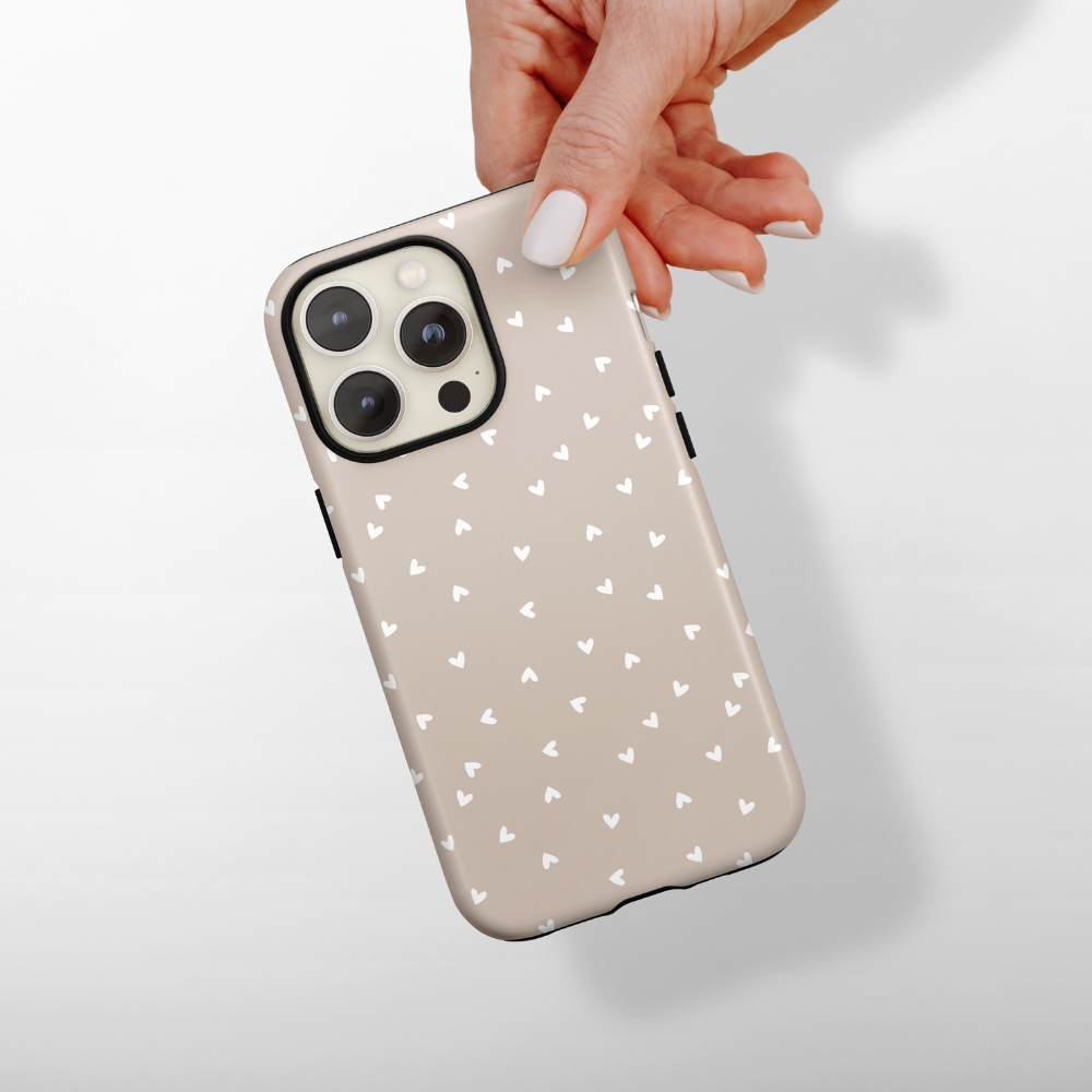 Hand holding a phone in a Coconut Lane Tough Phone Case with white hearts on a light chai-colored background.