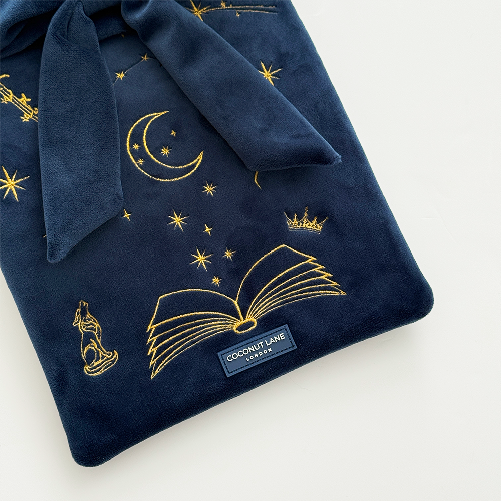 Book Sleeve made from soft navy blue velvet with gold embroidered crescent moon, stars, crown and open book illustrations, finished with a large velvet bow and Coconut Lane logo, photographed on a white background.