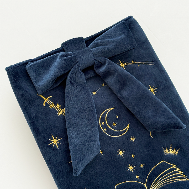 Book Sleeve made from soft navy blue velvet with gold embroidered crescent moon, stars, crown and open book illustrations, finished with a large velvet bow and Coconut Lane logo, photographed on a white background.