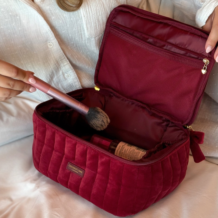 Burgundy velvet cosmetic bag with a quilted design, top handle, bow detail, and gold-tone hardware. Open showing makeu brush