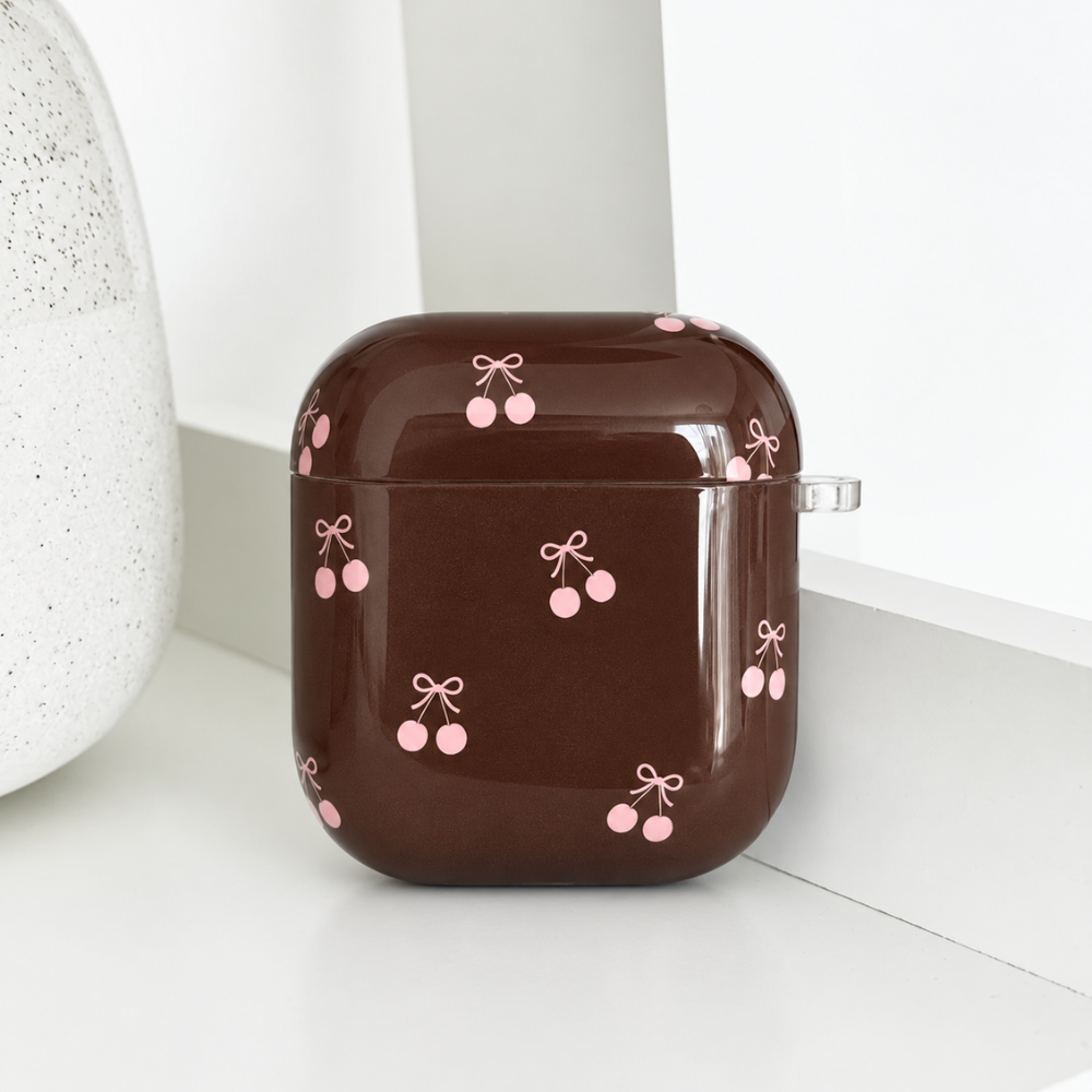 Glossy brown AirPods case with a pink cherry and bow print, closed and resting on a white shelf.