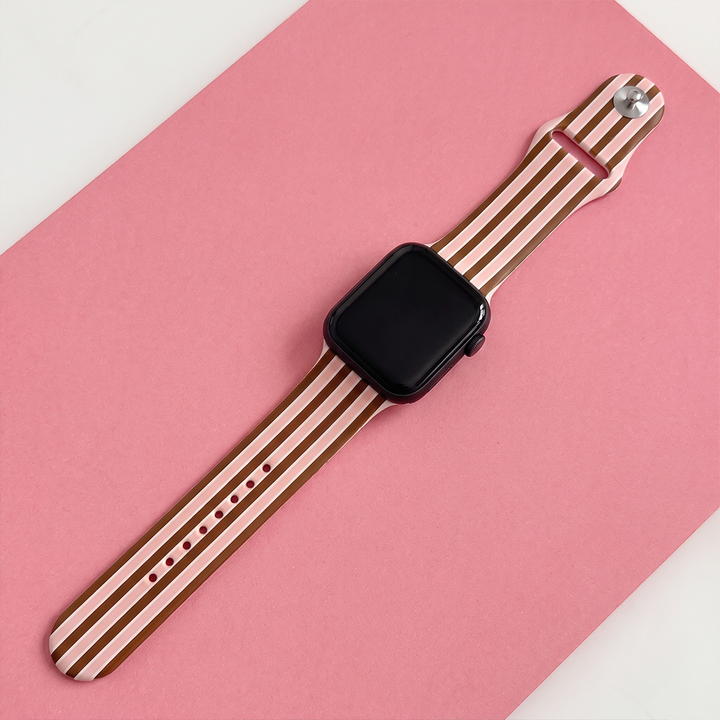 Apple Watch strap with brown, pink and cream stripes, displayed diagonally with a black Apple Watch on a pink background.