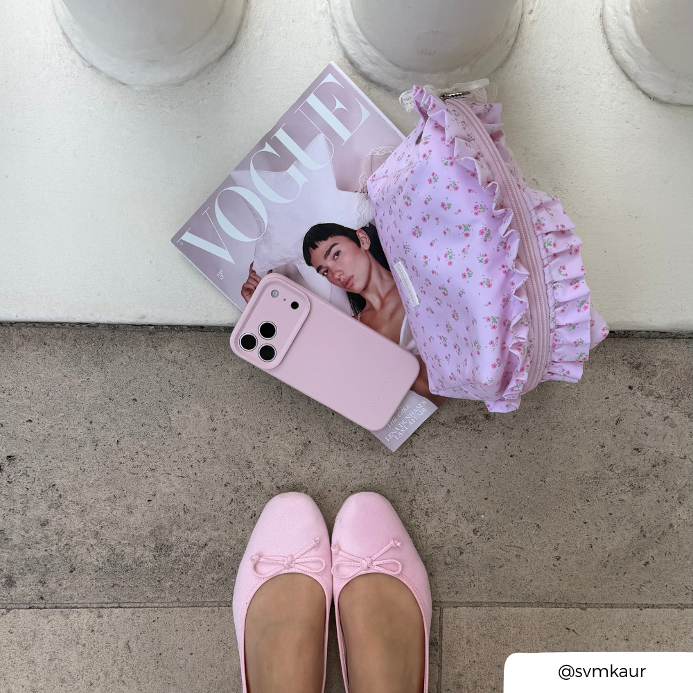 A styled flat-lay featuring a light pink floral ruffled makeup bag, a baby pink phone case, and a Vogue magazine on a concrete surface, with a person's feet in pink ballet flats visible at the bottom.