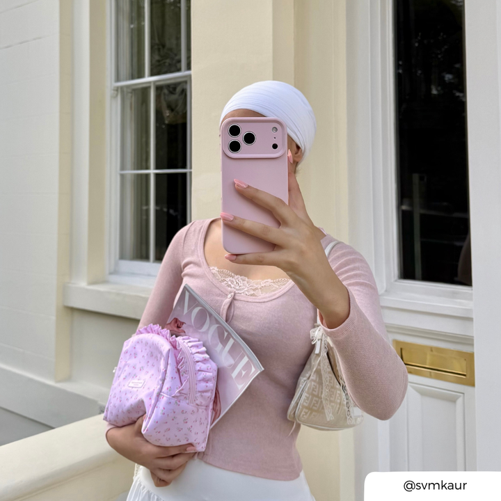 Woman in a baby pink long sleeve cardigan taking a selfie. She is holding an iphone in a baby pink phone case. In her hand she holds a ditsy floral pink makeup bag and a vogue magazine. 