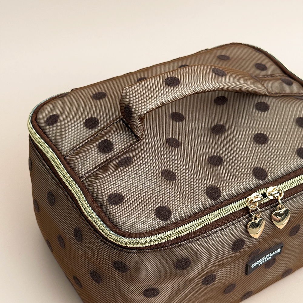 Birds eye view of brown mesh vanity case with dark brown polka dots, a padded top handle, gold zip with two heart-shaped zip pulls, and a black Coconut Lane London logo badge on the front, shown on a beige background.