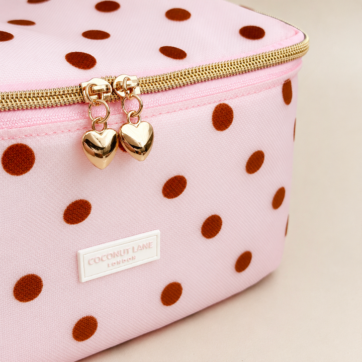 Close-up of pink mesh vanity case with brown polka dots, a padded polka dot top handle, gold zip with two heart-shaped zip pulls, and a white Coconut Lane London logo badge on the front, shown on a beige background.