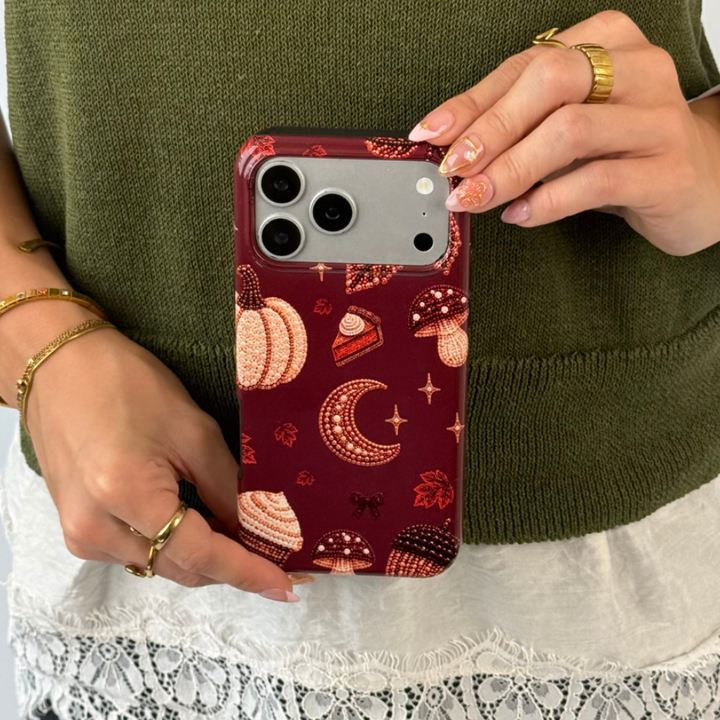 A person holding a phone with a dark red autumn-themed case featuring illustrations of pumpkins, a crescent moon, and a slice of pumpkin pie.