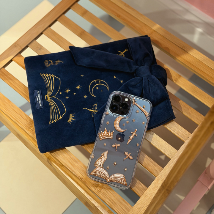 Clear fantasy girl phone case with golden illustrations of fantasy themed beaded effect icons, such as a book, wolf, crown, moon, sword, stars and dragons. With matching navy blue velvet book sleeve with matching design.