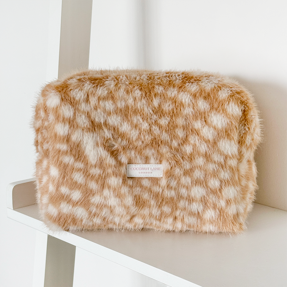 A fluffy faux fur cosmetic pouch in a tan and white spotted pattern, featuring a small white brand label that reads 'COCONUT LAUGH LONDON', resting on a white shelf.