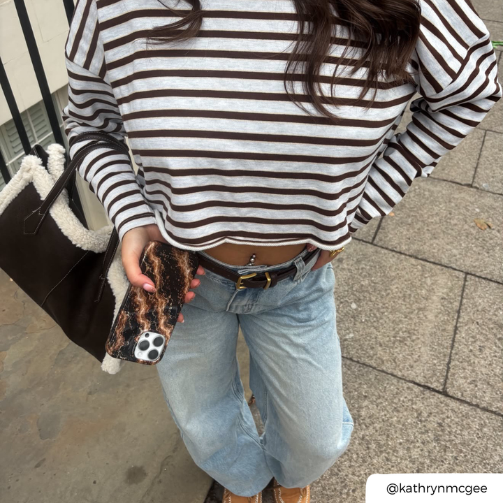 A woman with blue jeans and a striped long sleeve top, holds an iphone in a beige and dark brown marble design with subtle gold glitter flecks phone case
