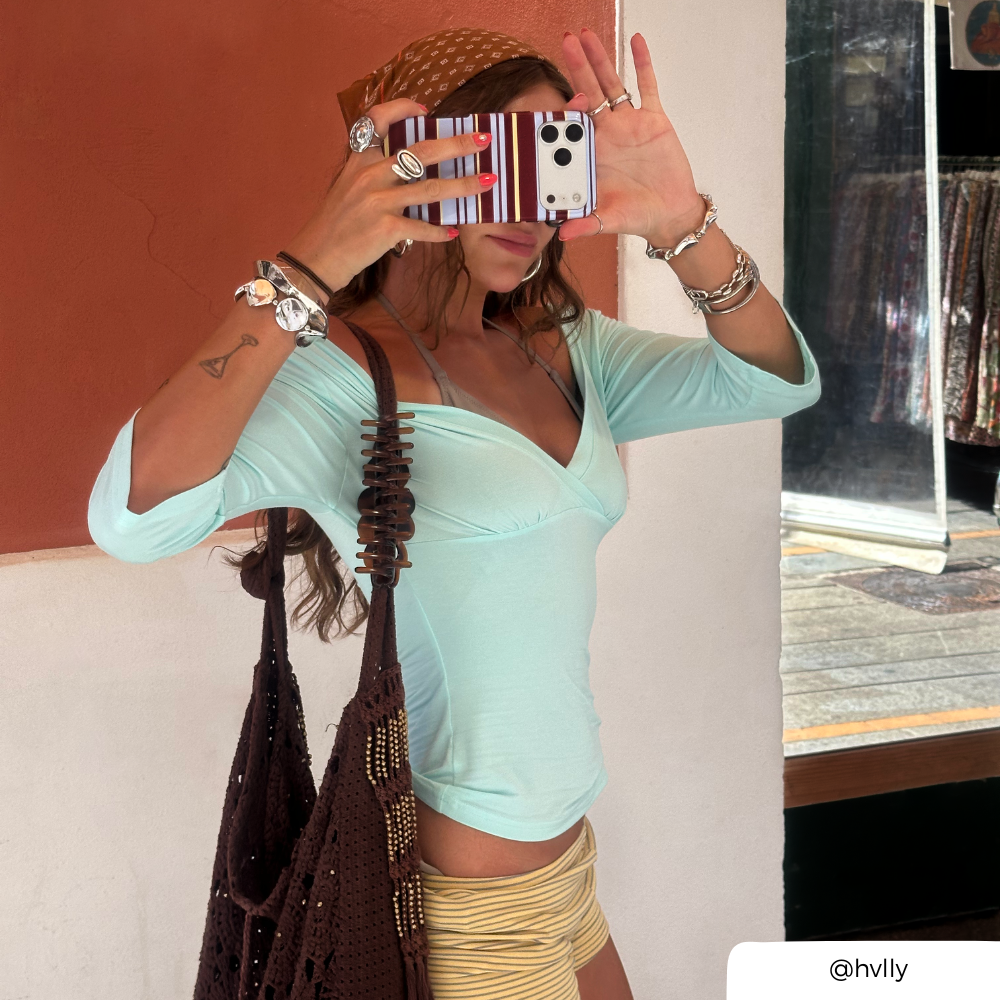 A woman takes a mirror selfie wearing a light blue green long-sleeve wrap top and a brown patterned headwrap, carrying a large brown knit shoulder bag. she is holding an iphone with a striped brown, blue and yellow phone case.