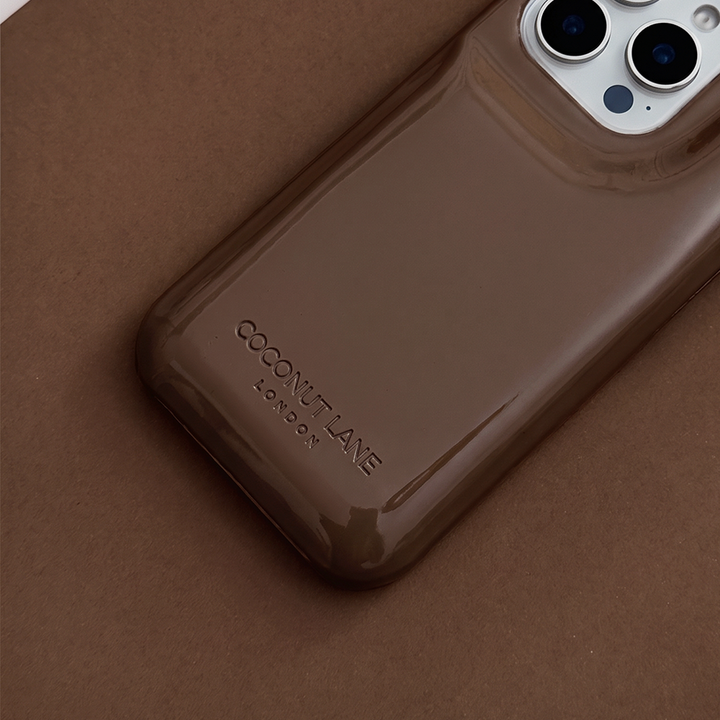 Close-up of glossy chocolate brown Coconut Lane puffer phone case with embossed logo, displayed diagonally on a matching brown background.
