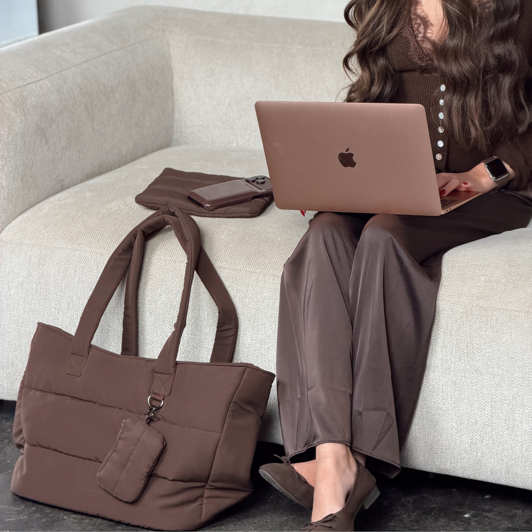 Woman with long brunette hair wearing a brown office outfit sat typing on a laptop. Next to her is the Main Carry Choc Brown Puffer bag, with the matching keyring bag attached on the d-ring. On the sofa next to her is the matching choc brown everything pouch and puffer phone case. 