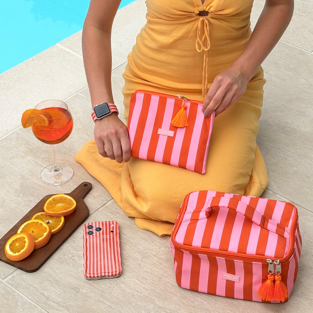 Person holding a pink and orange striped pouch next to a aperol and with matching phone case and vanity case