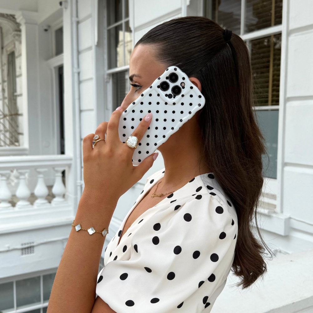 Woman holding a white polka dot wavy phone case to her ear outside a white building.