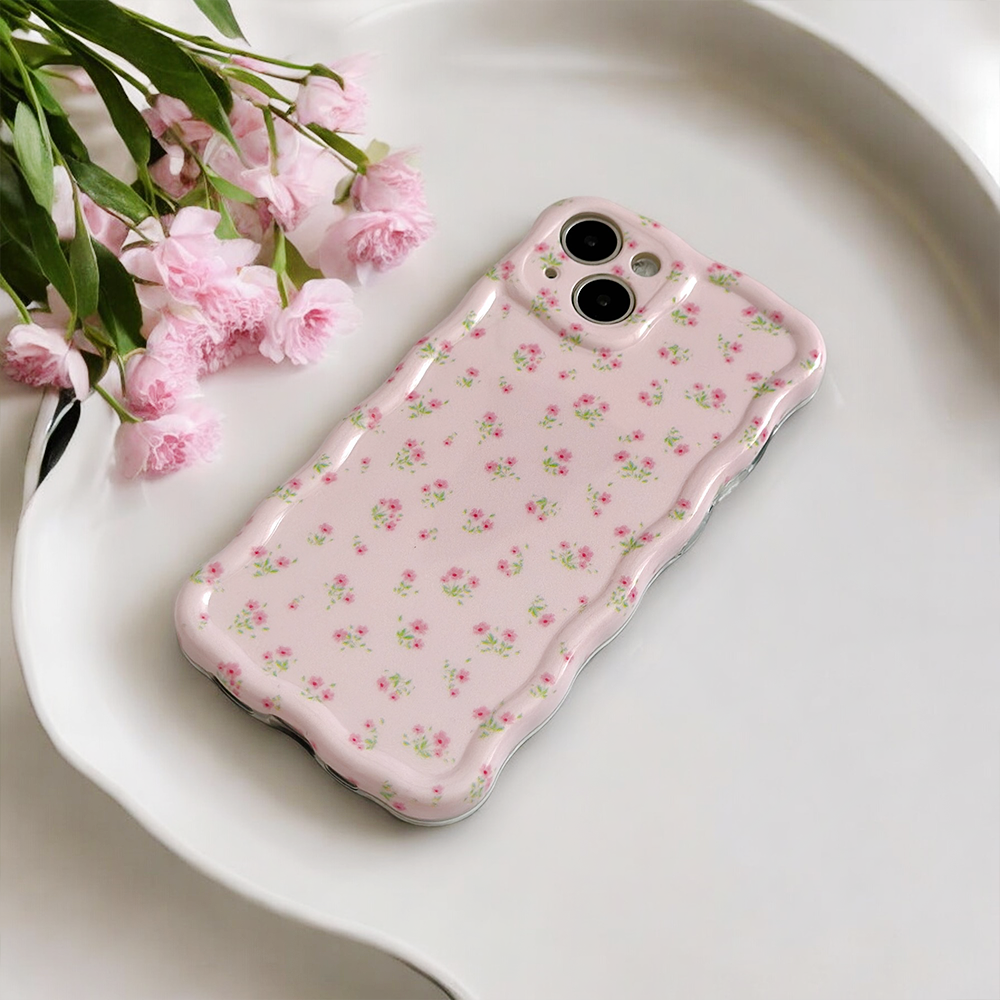 A pink smartphone with a Coconut Lane Wavy Phone Case - Ditsy Floral Pink, featuring small pink and green patterns, lies on a white tray. A bouquet of pink flowers rests beside the phone, adding a touch of elegance to the composition.