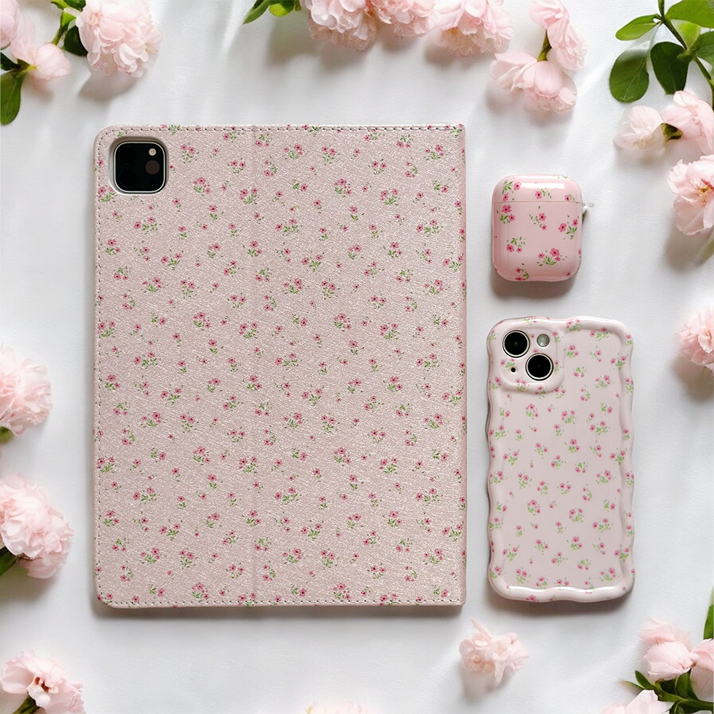 A tablet, a phone protected by Coconut Lane's Wavy Phone Case in Ditsy Floral Pink, and an AirPods case all featuring matching pink floral designs are arranged on a white surface surrounded by pale pink blossoms. The cases showcase a delicate pattern of ditsy floral pink flowers, creating a cohesive and aesthetically pleasing look.