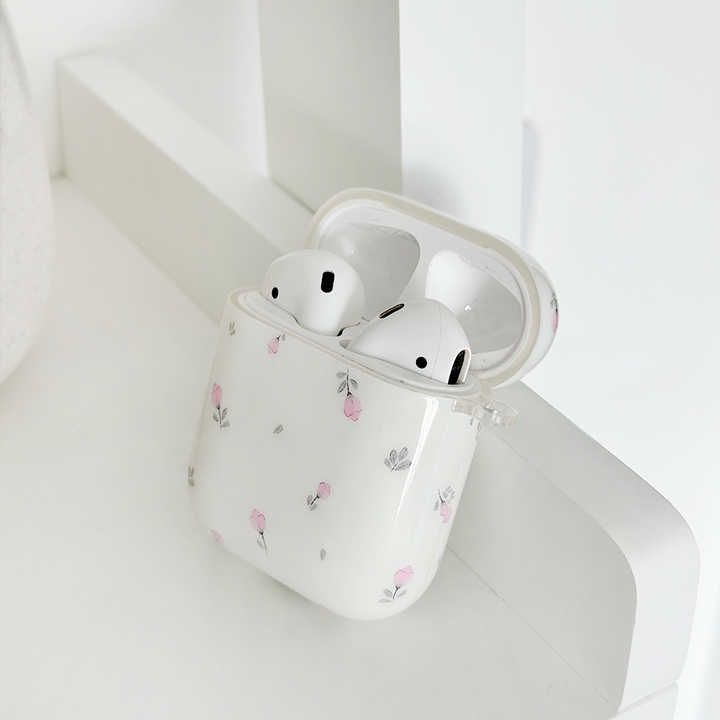 A Coconut Lane Airpods Case - Ditsy Floral White, featuring a delicate floral pattern, rests on a white surface. The lid is open, revealing two white earbuds inside. The overall aesthetic is minimalistic and clean, with soft pink and gray floral accents on the protective cover.