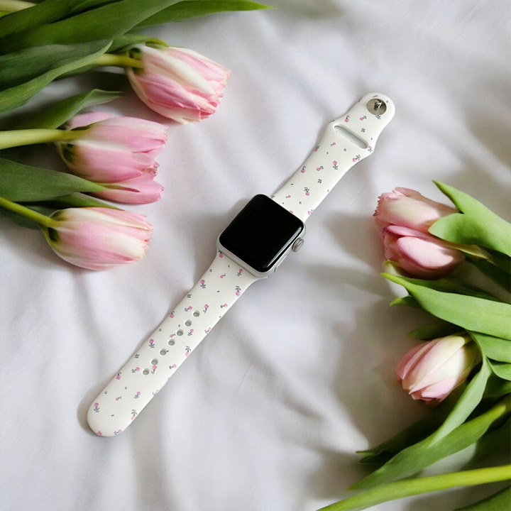 A smartwatch featuring a Ditsy Floral White Apple Watch Strap from Coconut Lane is placed on a white surface, surrounded by pink tulips with green leaves. The flowers are arranged artistically around the watch, creating a fresh, spring-like scene.