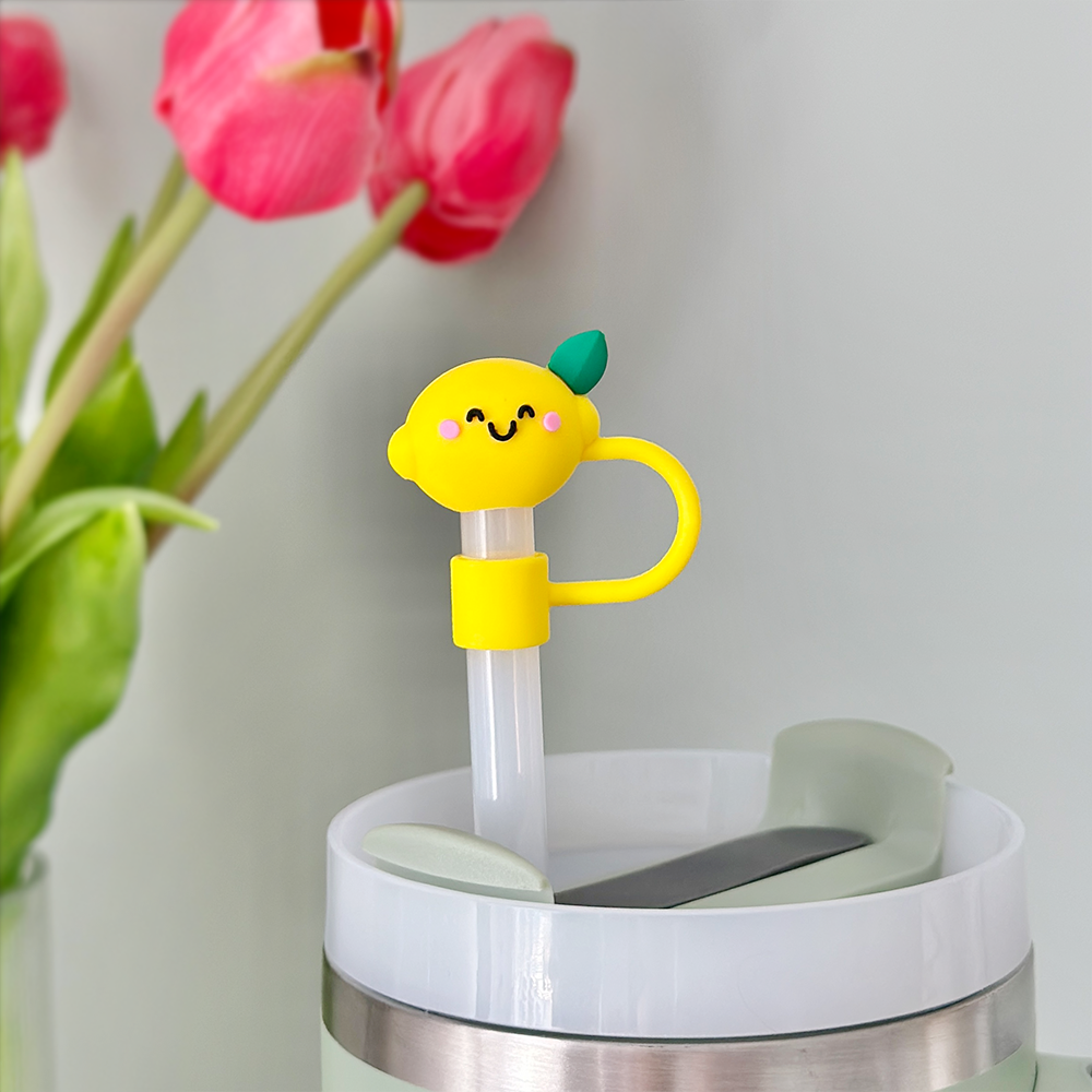 A close-up of a drink cup lid with a reusable straw featuring Coconut Lane's Happy Lemon Straw Cover, which is made of high-quality silicone and includes a lemon-shaped top with a smiling face and green leaf. In the background, soft-focused pink tulips are arranged in a vase.