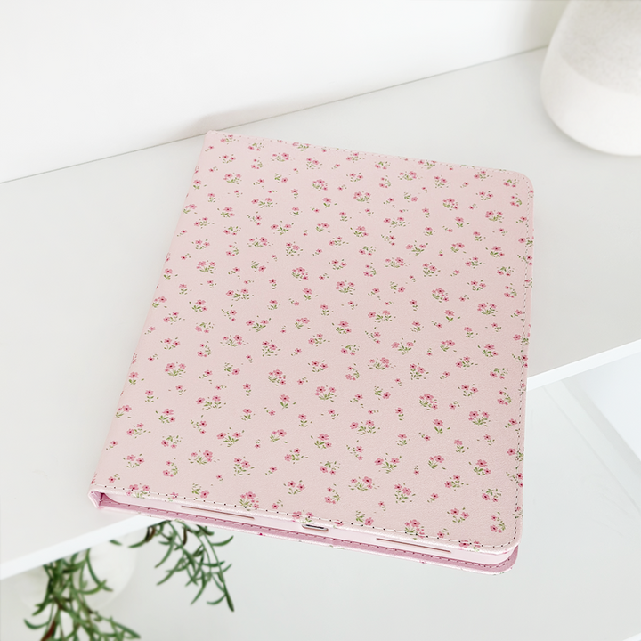 A pink notebook adorned with a delicate floral pattern rests on a white surface. A plant with lush green leaves is partially visible in the corner, and to the right, a white cylindrical object, likely a vase, is seen in the background. The setting perfectly complements my Coconut Lane Ditsy Floral Pink iPad Case.