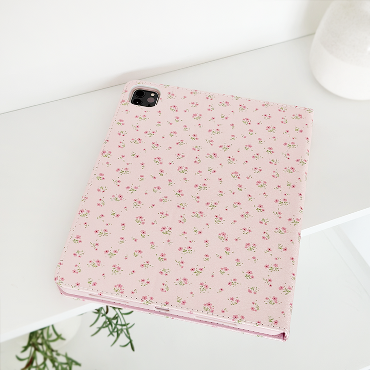 A Ditsy Floral Pink iPad Case by Coconut Lane lies on a white shelf. This vegan leather case, decorated with delicate floral patterns, features a camera cutout in the top left corner. The shelf also holds a white vase and a small plant, adding a touch of greenery to the minimalist setting.