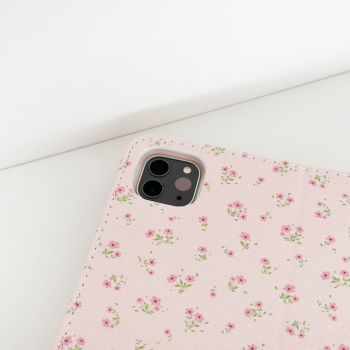 A corner of a smartphone with a Coconut Lane Ditsy Floral Pink iPad Case, showcasing small, delicate flowers in shades of light pink and green, is placed on a white surface. The phone's three rear cameras and flash are visible. This design is also available for the Apple iPad case collection.