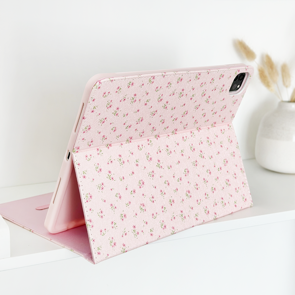 A tablet with a Coconut Lane Ditsy Floral Pink iPad Case is propped up on a white surface. The case functions as a stand, holding the tablet at an angle. In the background, there is a white ceramic vase with dried grasses for decoration.