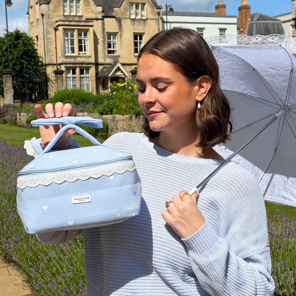 Woman holding blue vanity case with lace details and an umbrella in a garden setting.