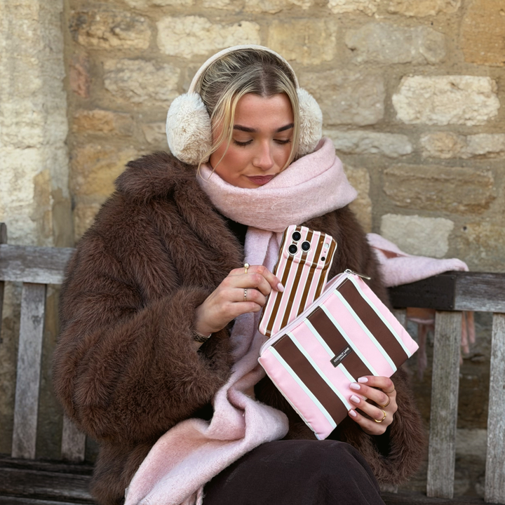 Woman wearing a baby pink scarf and faux fur brown jacket with cream fluffy ear muffs on her head. She is holding an iphone in a cocoa & Pink Stripe Wavy Phone Case featuring glossy brown, blush pink and cream vertical stripes, photographed diagonally on a pink background. In her hand is the matching Cocoa and Pink stripes everything pouch. 