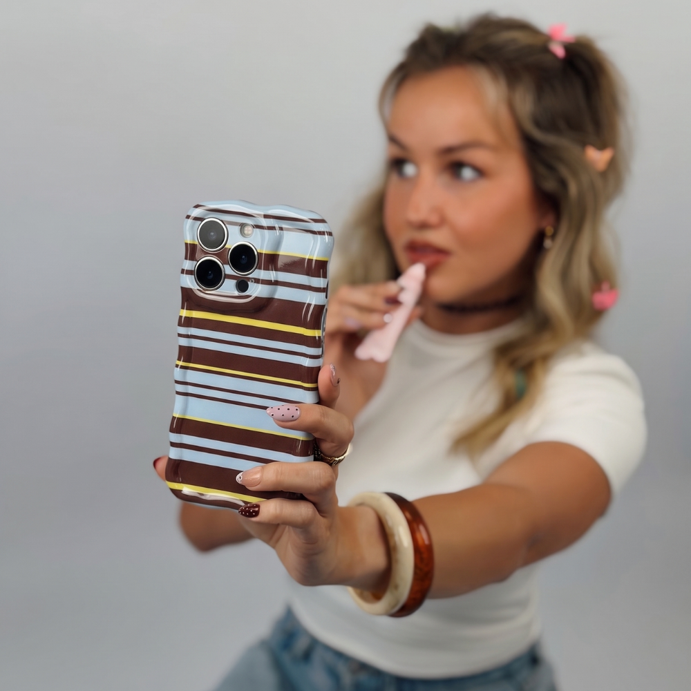 girl holding a wavy brown, blue and yellow phone case. Wearing a white t-shirt and jeans. Applying lip gloss