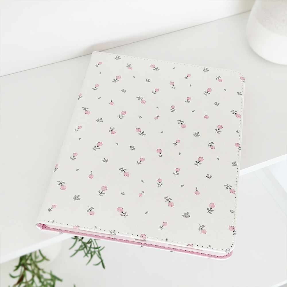 A white iPad case with a delicate pink and grey floral pattern from Coconut Lane, named the Ditsy Floral White iPad Case, sits on a white surface. The surrounding space is minimalistic, featuring a small green plant peeking in from the bottom left corner and a textured white object in the top right. It's as elegant as an anti-slip Vegan Leather accessory.