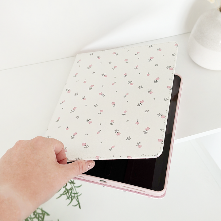 A hand is holding a Coconut Lane Ditsy Floral White iPad Case, partially opening it to reveal a black tablet inside. The vegan leather cover is white with small pink flowers. The scene is set against a white background with a small green plant and a white vase visible in the frame.