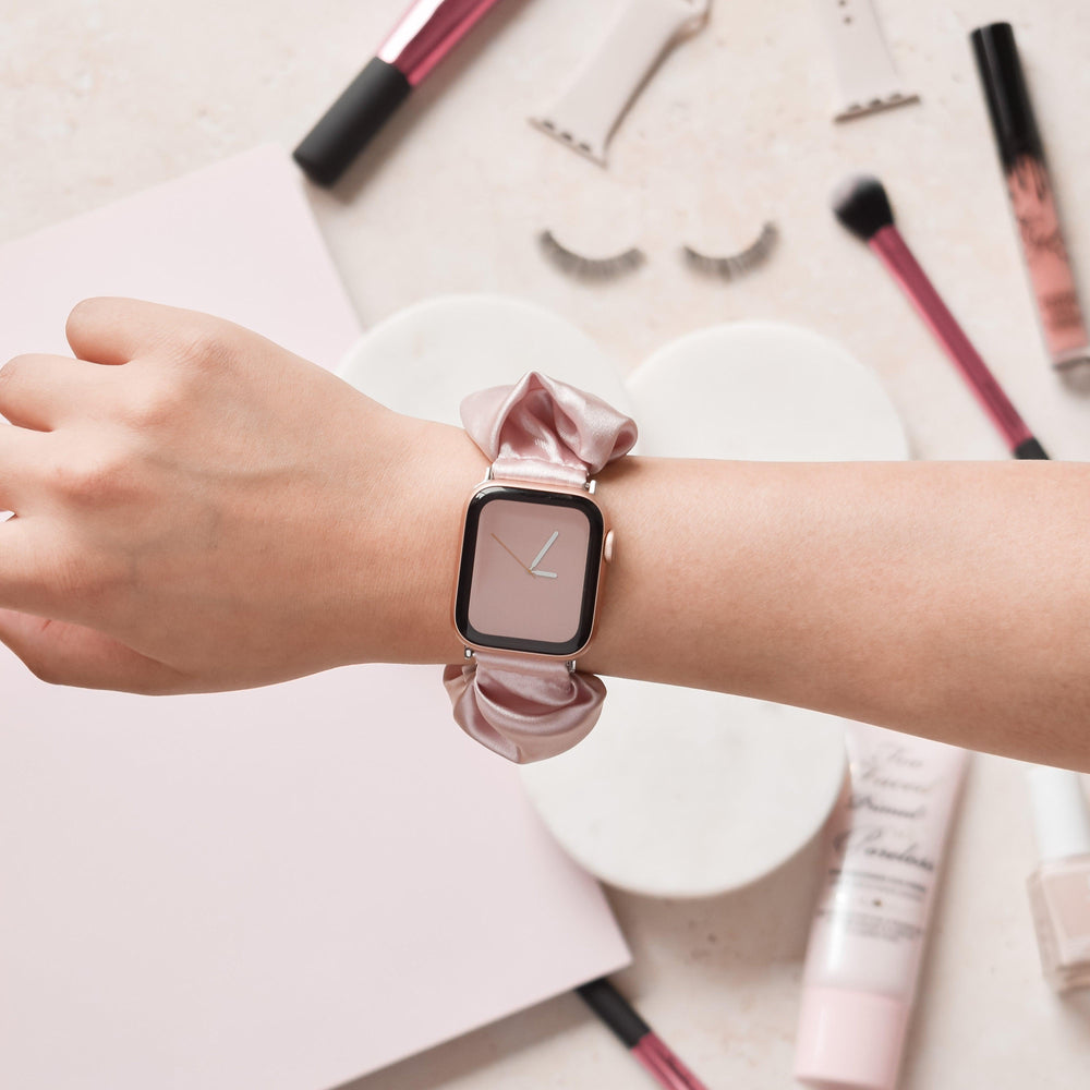 Hand wearing a powder pink Coconut Lane scrunchie-style Apple Watch strap near assorted makeup items on a table.